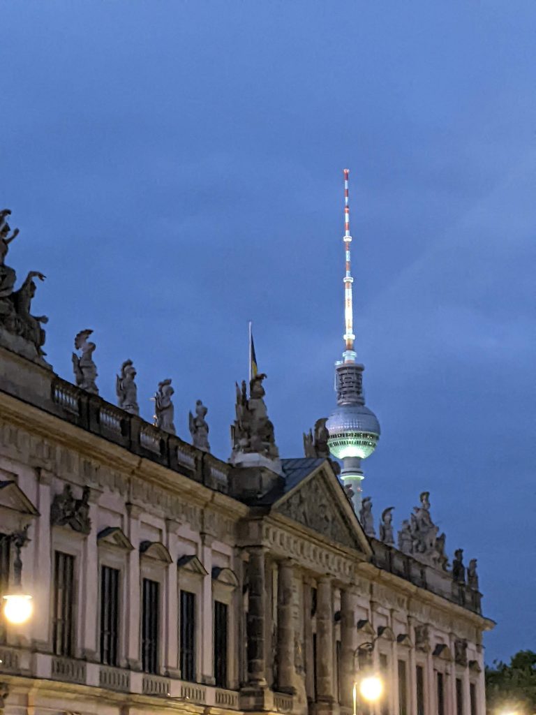 Nächtliche Aufnahme vom Fernsehturm in Berlin. Davor eine historische Fassade mit Giebelfiguren. 