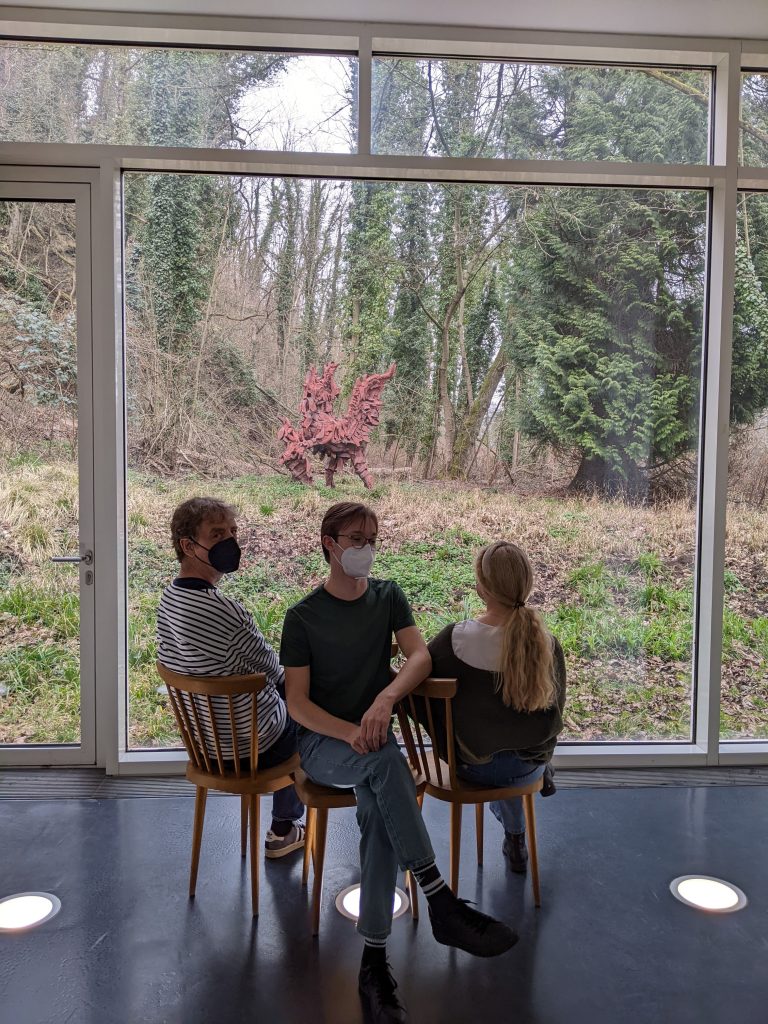 Drei Menschen sitzen gegeneinander versetzt auf einem Dreiermöbel und blicken in den Garten durch eine große Glasscheibe. Sie haben Corona-Schutz-Masken auf. 