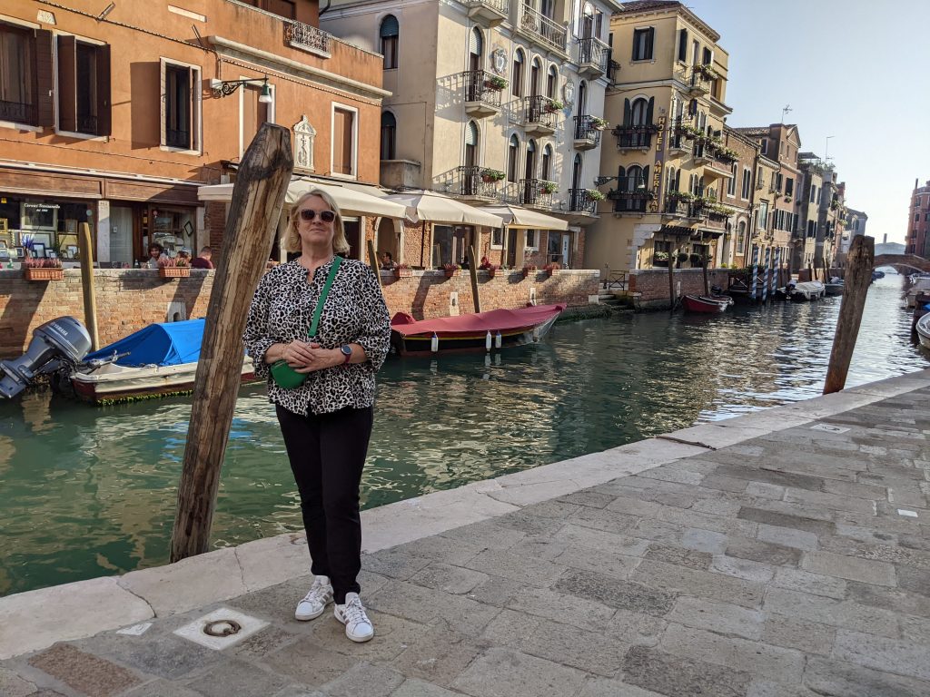 Frau in getigertem Oberteil mit schwarzer Hose vor einem Kanal in Venedig. Im Hintergrund Boote. 