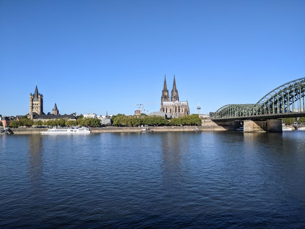 Panorama der Stadt Köln. Mit der Hohenzollernbrücke am rechten Bildrand. Links daneben der Kölner Dom. Etwas weiter links Groß St. Martin, eine der romanischen Kirchen Kölns. Blauer Himmel. Im Vordergrund der Rhein. 