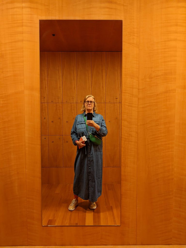 Frau mit blauem Jeanskleid macht ein Spiegelselfie. Im Hintergrund Museumsschließfächer in Holzoptik. 